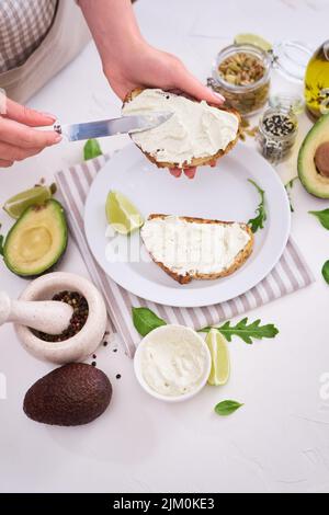 Zubereitung von Avocado- und Frischkäse-Toasts - Frau schmiert Käse auf einem gegrillten oder getoasteten Brot Stockfoto