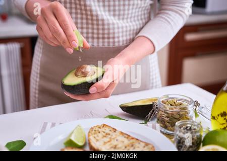 Frau quetscht frischen Limettensaft auf einer halbierten Avocado in der heimischen Küche Stockfoto