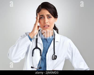 Wie kann ich meinen Patienten mit diesen schrecklichen Kopfschmerzen helfen. Studioaufnahme eines jungen Arztes, der vor grauem Hintergrund Stress erlebt. Stockfoto