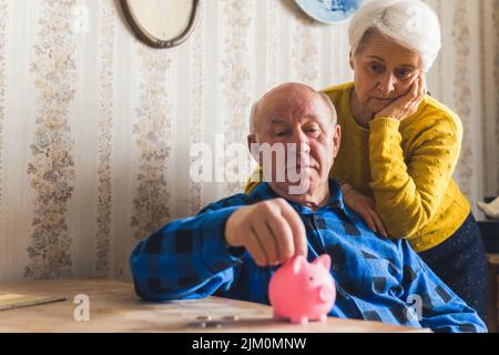 Trauriges, verlorenes und besorgter kaukasischer Senioren, der Münzen zählte, die von einem Sparschwein genommen wurden, während er am Tisch saß. Finanzkrise. Hochwertige Fotos Stockfoto
