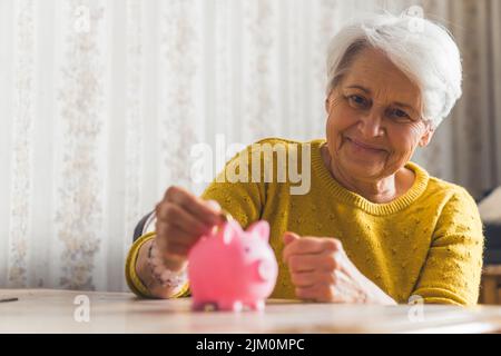 Optimistische, fröhlich lächelnde kaukasische ältere Frau, die Münzen in ein Sparschwein legt. Einsparungen. Hochwertige Fotos Stockfoto