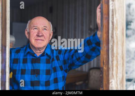 Kahlköpfiger, gutaussehender kaukasischer älterer Mann, der aus dem offenen Fenster schaute, wobei eine seiner Hände an der Wand ruhte. Hochwertige Fotos Stockfoto