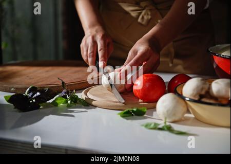 Hände eines Küchenchefs, der frische Champignons auf einem hölzernen Schneidebrett in der Nähe von Bio-Gemüse und kulinarischen Kräutern hackt Stockfoto