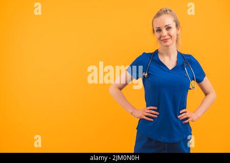 Junge blonde, weiße Krankenschwester mit Pferdeschwanz und Scrubs, die mit lächelnden Händen auf den Hüften stehen und in die Kamera blicken. Studioaufnahme. Hochwertige Fotos Stockfoto
