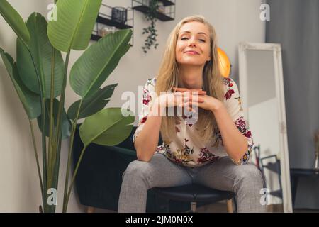 Blonde weiße junge Frau im Wohnzimmer, die die Arme auf den Knien in selbstbewusster Pose aufliegt, den Kopf hoch über den Händen und lächelnd gehalten. Innenaufnahme. Hochwertige Fotos Stockfoto