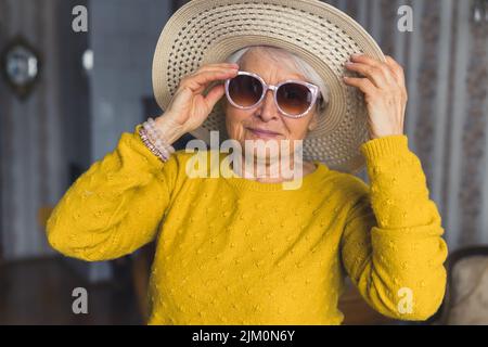 Lustige kaukasische ältere alleinerziehende Frau, die eine stilvolle Sonnenbrille und einen Strandhut trägt, Spaß im Ruhestand hat und sich für den Urlaub vorbereitet. Hochwertige Fotos Stockfoto