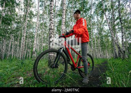 Eine Radfahrerin fährt auf der Straße in einem wunderschönen dichten grünen Wald mit dem Rennrad. Bikegirl in Mystery Forest. Stockfoto
