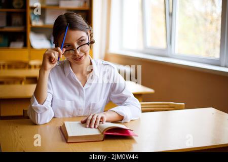 Eine fokussierte, tausendjährige Schülerin sitzt im Klassenzimmer und studiert mit dem Schreiben von Referenzbüchern. Machen Sie Hausaufgaben, konzentriert Teenager-Mädchen nimmt Notizen in Notizbuch Stockfoto