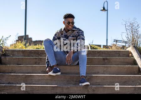 Porträt eines lächelnden jungen lateiners mit Sonnenbrille, der im Sommer auf der Straße sitzt Stockfoto