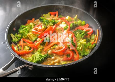 Dampfendes Gemüsekry mit Kichererbsen, Brokkoli und rotem Paprika in Kokosmilch in einer Pfanne auf einem schwarzen Herd, gesunde Mahlzeiten für einen veganen Low Stockfoto