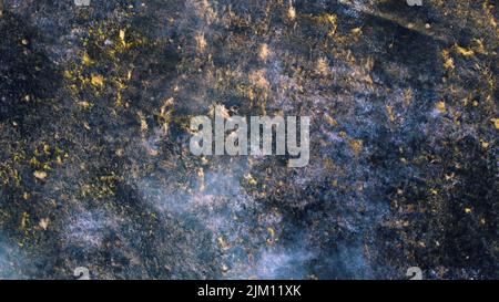 Luftdrohnen-Rundflug über schwarzer Aschefelde aus verbranntem braungelbem, trockenem Gras und weißem Rauch am sonnigen Herbsttag. Blick von oben. Textur natürlicher Hintergrund der ökologischen Umweltkatastrophe. Stockfoto