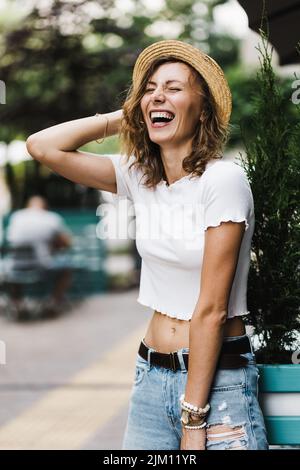 Optimistische Frau mit lockigen Haaren in Jeans lacht aufrichtig in der Stadt. Eine edle Dame in weißer Bluse und schließt ihre Augen vor Glück. Stockfoto