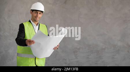 Junger Architekt Mann trägt Auftragnehmer Helm hält Projekt Papier Plan über isolierten Hintergrund glücklich mit großem Lächeln, Wand der Konstruktion Stockfoto
