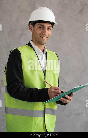 Muslimischer junger Ingenieur, der auf der Zwischenablage Notiz nimmt, Porträt eines Ingenieurs in einem weißen Helm, männlicher Architekt, der auf der Zwischenablage schreibt Stockfoto