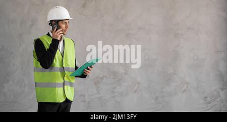 Männlicher Architekt, der die Architekturzeichnung auf der Baustelle überprüft. Mann in der Uniform des Baumeisters benutzt Telefon, während er Blaupausen liest. Anrufe von Architekten Stockfoto