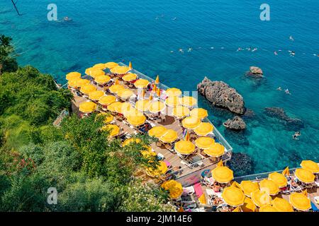23 June 2022, Antalya, Türkei: Versteckter, enger und gemütlicher Strand zwischen Klippen und Felsen mit leuchtend gelben Sonnenschirmen und Sonnenliegen und landschaftlich erfrischendem Blau Stockfoto