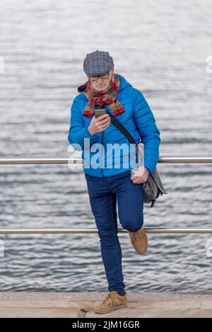 Älterer Mann mit Brille, der sich am Meer am Geländer lehnt, während er sein Smartphone benutzt Stockfoto