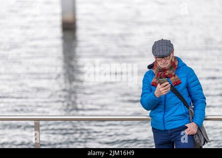 Älterer Mann mit Brille, der sich am Meer am Geländer lehnt, während er sein Smartphone benutzt Stockfoto