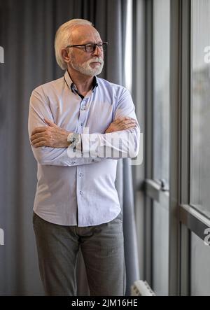 Selbstbewusster Geschäftsmann, der durch das Fenster schaut. Der männliche Profi steht mit gekreuzten Armen. Er trägt im Büro der Unternehmensleitung Formalien. Stockfoto