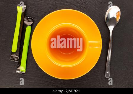 Zwei Zuckerstäbchen in Papiertüten mit einer Keramikuntertasse, einer Tasse und einem Metalllöffel auf einem Schieferstein, Nahaufnahme, Draufsicht. Stockfoto