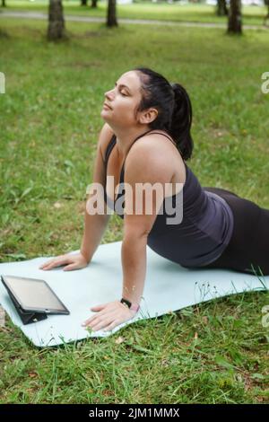 Eine charmante Brünette Frau plus-Größe Körper positiv praktiziert Sport in der Natur. Frau macht Yoga im Park auf einer Sportmatte Stockfoto