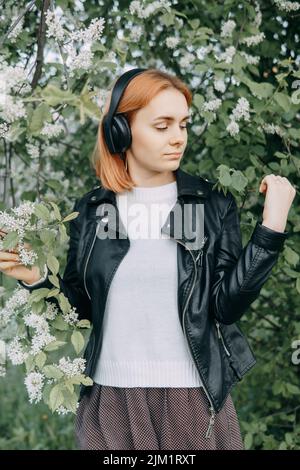 Ein rothaariges Teenager-Mädchen hört Musik über Kopfhörer, die über das Telefon konfiguriert werden. Die Zeit der Frühjahrsblüte der Kirschgärten Stockfoto