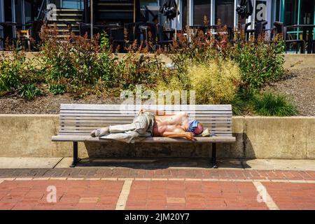 Baltimore, MD, USA – 2. August 2022: Eine Obdachlose schläft tagsüber auf einer Bank im Binnenhafen von Baltimore in der Innenstadt. Stockfoto