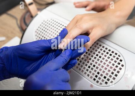 Der Manikurist schneidet die Nagelhaut mit einer kleinen Schere und trägt blaue Latexhandschuhe. Stockfoto