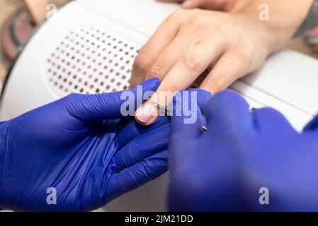 Der Manikurist schneidet die Nagelhaut mit einer kleinen Schere und trägt blaue Latexhandschuhe. Stockfoto