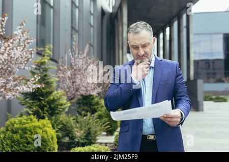 Leitender und erfahrener grauhaariger Chef-Mann in der Nähe des Büros nachdenklich beim Lesen des Finanzberichts Dokument außerhalb des Büros, Geschäftsmann denken Stockfoto