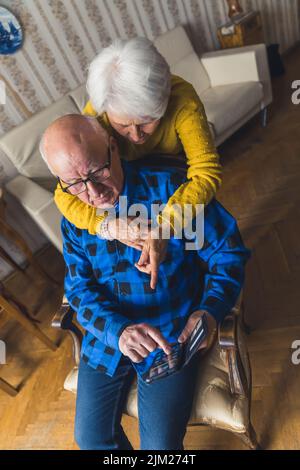 Kaukasische Senior verwirrt Paar versucht zu lernen, wie man ein Smartphone zu verwenden. Ein Mann, der auf einem Stuhl sitzt, während eine Frau ihn von hinten umarmt und auf den Bildschirm zeigt. Hochwertige Fotos Stockfoto