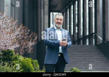Porträt eines erfolgreichen grauhaarigen Senior-Bankiers vor dem Bürogebäude, lächelnder Mann mit Telefon und Blick auf die Kamera, glücklicher Geschäftsmann im Geschäftsanzug Stockfoto