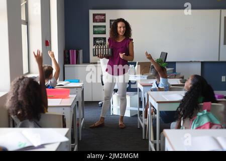 Kaukasische junge Lehrerin mit Blick auf multirassische Grundschüler, die während des Unterrichts die Hand heben Stockfoto