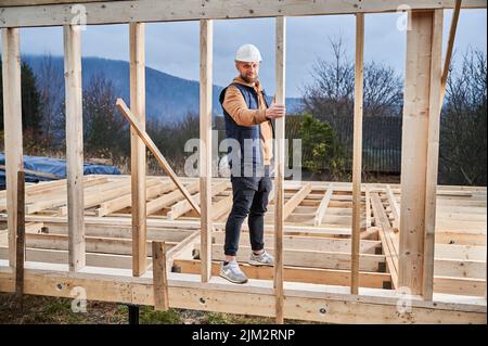Männlicher Baumeister Gebäude Holzrahmenhaus. Porträt eines Mannes, der auf der Baustelle in einem Schutzhelm steht und die Qualität der Arbeit überprüft. Stockfoto