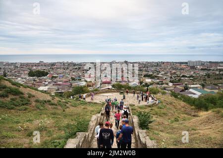 Derbent, Russland - 24. Juli 2022: Touristen gehen von der Festung Naryn-Kala auf die Treppe hinunter. Moderne Stadt Derbent und Kaspisches Meer auf dem Hintergrund. Stockfoto