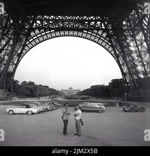 1961, historische, Autos aus der Zeit, die unter dem Eiffelturm in Paris, Frankreich, geparkt wurden, wobei zwei Männer auf dem offenen Gelände unter der riesigen Metallstruktur standen, die für die Pariser Ausstellung 1889 gebaut wurde. Ursprünglich als provisorisches Bauwerk gedacht, sollte der schmiedeeiserne Turm 1909 abgerissen werden, wurde aber aufgrund seiner Verwendung als Radiotelestation vor der Zerstörung bewahrt. Es ist heute eines der berühmtesten Wahrzeichen der Welt. Stockfoto