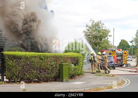 Rauch und Flammen wehend als Gruppe von Feuerwehrmänner zielen Wasserstrahl an Hausfront und Seitenwänden Essex Fire & Rescue Tender Wohnstraße Essex England UK Stockfoto