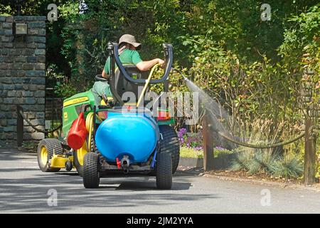 Gärtner, der hoch oben auf dem John Deere Minitraktor sitzt und einen blauen Mini-Pumpwassertanker schleppt, der Blumen im Landgut des Hotels England versprüht Stockfoto