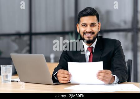 Positive gutaussehende selbstbewusste arabische oder indische erfolgreiche Geschäftsmann, Anwalt, Assistent, in einem formellen Anzug, sitzen am Schreibtisch mit Laptop in modernen Büro, hält Dokument, schaut auf die Kamera, lächelnd Stockfoto