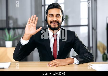 Freundlicher, eleganter indischer oder arabischer Geschäftsmann, Finanzberater, sitzen im modernen Büro am Schreibtisch, führen Videogespräche mit dem Kunden, schreiben Online-Schulungskurs von männlichen Wirtschaftsexperten auf, lächeln Stockfoto