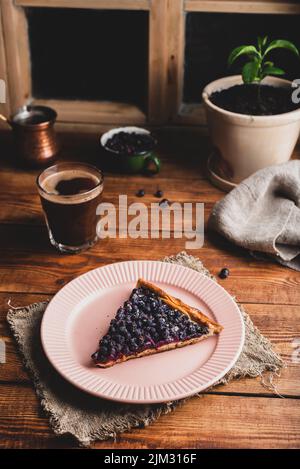 Ein Stück frisch hausgemachter Amelanchierbeeren auf dem Teller und ein Glas Kaffee auf dem rustikalen Tisch Stockfoto