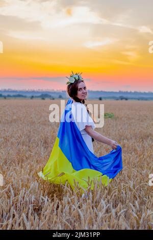Glückliches Mädchen in nationalen ukrainischen bestickten Hemd mit Kranz auf dem Kopf halten große gelb-blaue Flagge vor dem Hintergrund von Ädernweizen Feld. Stockfoto