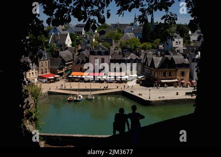 Ein Paar, das gegen den grünen Fluss Loch stand, blickt auf den Place Saint-Sauveur im Saint-Goustan-Bezirk Auray, Morbihan, im Süden der Bretagne, Frankreich, gesäumt von Restaurants und Straßencafés mit bunten Markisen und Sonnenschirmen. Saint-Goustan, einst ein geschäftiges Fischerei- und Handelshafen, ist heute ein beliebtes Touristenziel, das einem Besuch des US-Gründervaters Benjamin Franklin im Jahr 1776 Tribut zollt. Stockfoto