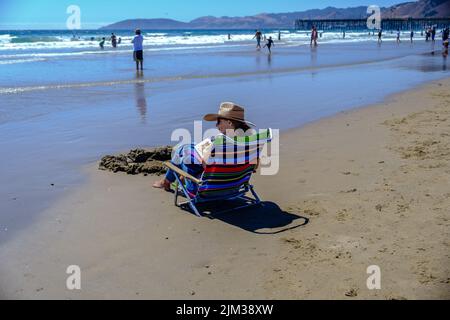 Lady in Sonnenhut sitzt auf einem Stuhl am Ufer und liest ein Buch am Pismo Beach, Kalifornien, mit Bergen und Pismo Pier im Hintergrund. Stockfoto