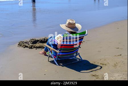 Lady in Sonnenhut sitzt auf einem Stuhl am Ufer und liest ein Buch am Pismo Beach, Zentralkalifornien. Stockfoto