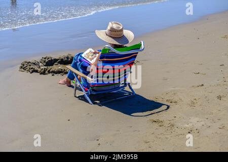 Lady in Sonnenhut sitzt auf einem Stuhl am Ufer und liest ein Buch am Pismo Beach, Zentralkalifornien. Stockfoto