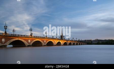 Pont de Pierre erstreckt sich über den Fluss Garonne in der Stadt Bordeaux und wird von der untergehenden Sonne mit der Basilique Saint-Michel im Hintergrund beleuchtet Stockfoto