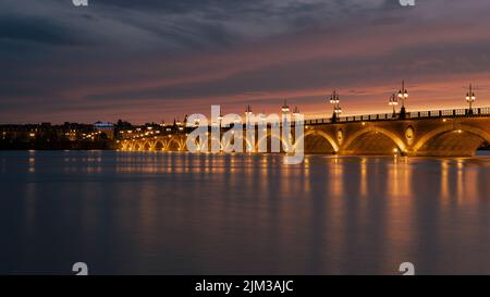 Pont de Pierre, der den Fluss Garonne in der Stadt Bordeaux überspannt, beleuchtet bei Sonnenuntergang mit der Basilique Saint-Michel im Hintergrund Stockfoto