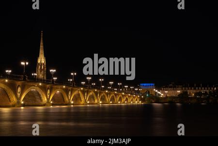 Pont de Pierre, der den Fluss Garonne in der Stadt Bordeaux überspannt, beleuchtet bei Nacht mit der Basilique Saint-Michel im Hintergrund Stockfoto
