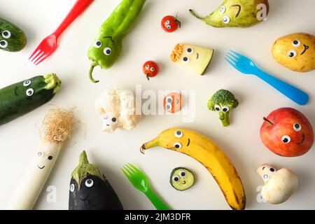 Hintergrund mit einer Vielzahl von lustigen Früchten und Gemüse präsentiert für die Kinderernährung mit Augen und Mund auf einem weißen Tisch mit farbigen Gabeln. Stockfoto